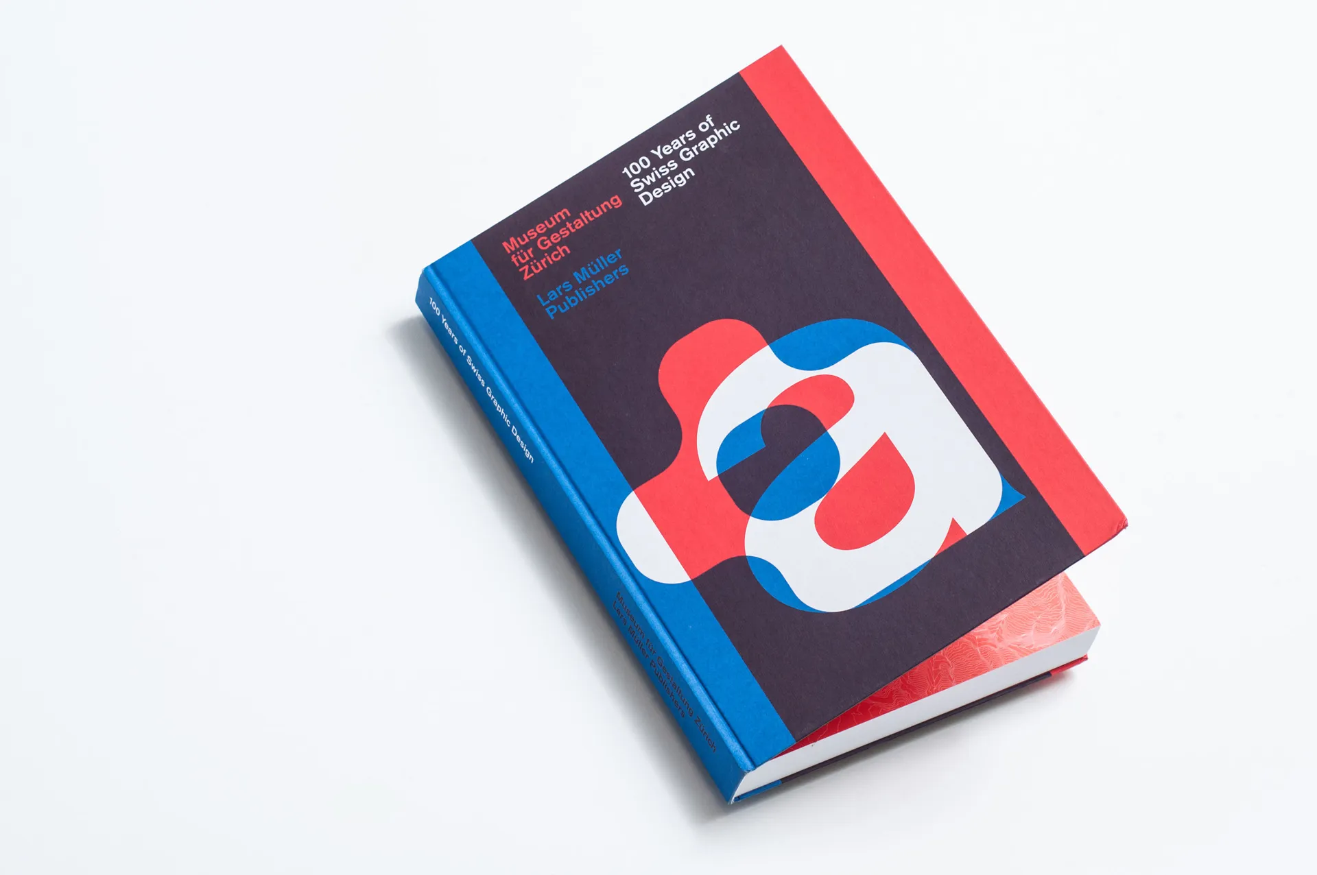 A book titled "Museum für Gestaltung Zürich: 100 Years of Swiss Graphic Design" with a blue and red cover. The front features a large, abstract design comprising of overlapping red, white, and blue shapes resembling the letter "a". The book rests on a whi
