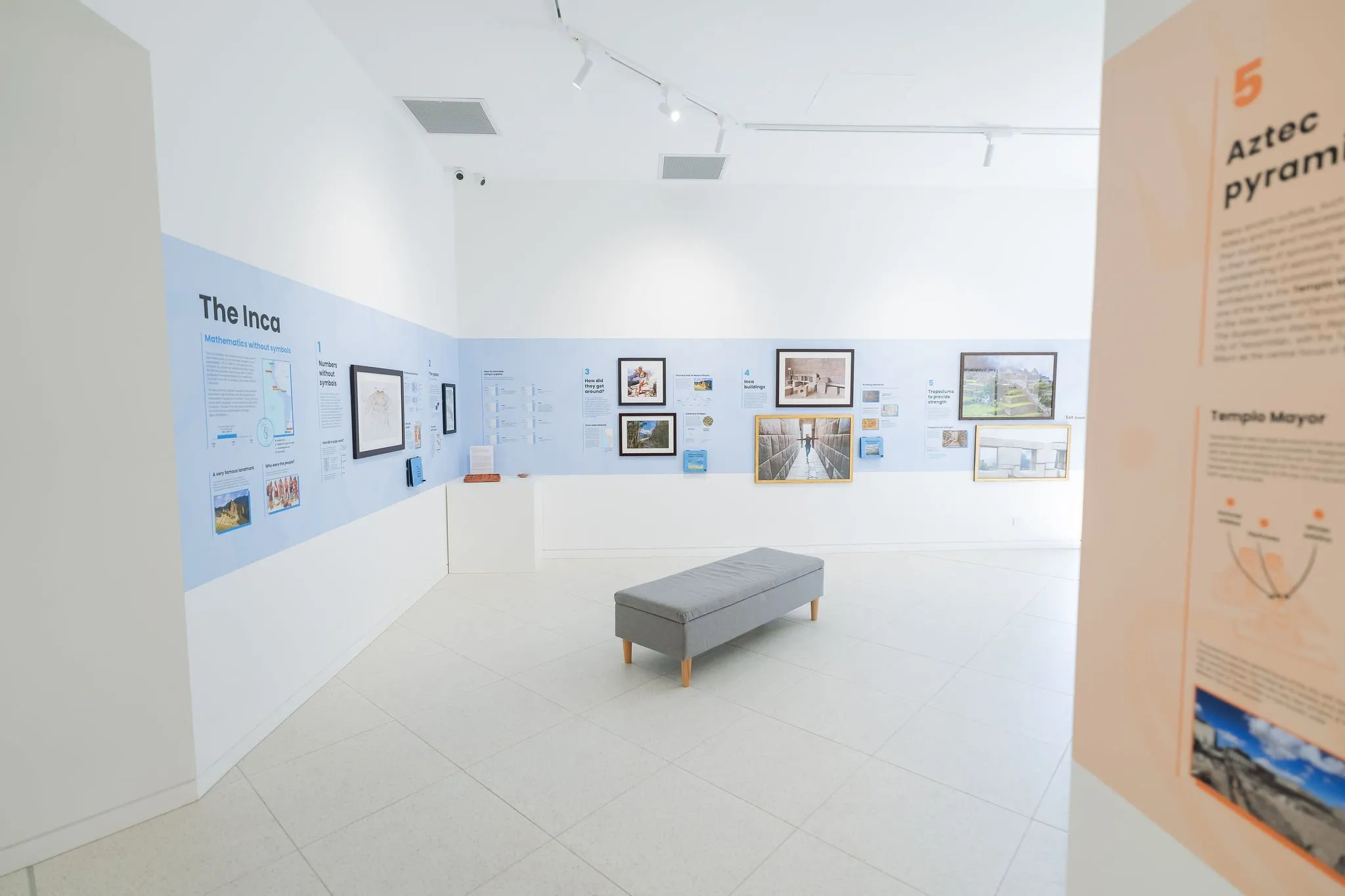 A modern exhibition room with white walls displays information and images about the Inca and Aztec civilizations. A central grey bench provides seating. Informational panels and pictures line the walls, with one prominently labeled "5 Aztec Pyramids.