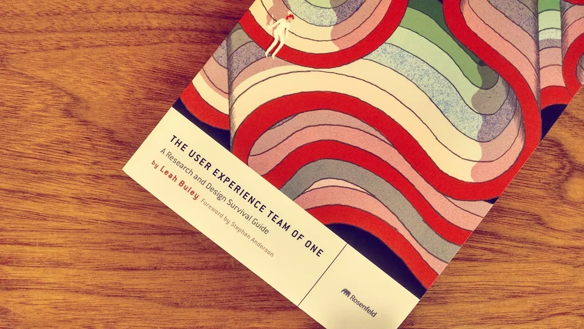 The image shows the book "The User Experience Team of One" by Leah Buley on a wooden surface. The book cover features a colorful abstract design with wavy lines in shades of red, pink, green, purple, and white. The author's name is at the bottom.