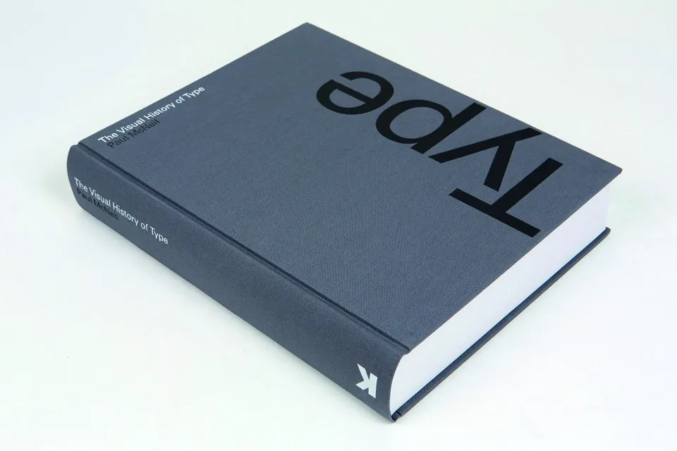 A hardcover book titled "The Visual History of Type" lies on a flat surface with its cover visible. The title is written in white and large black letters, positioned vertically and upside-down. The book has a grayish cover and appears to be thick.